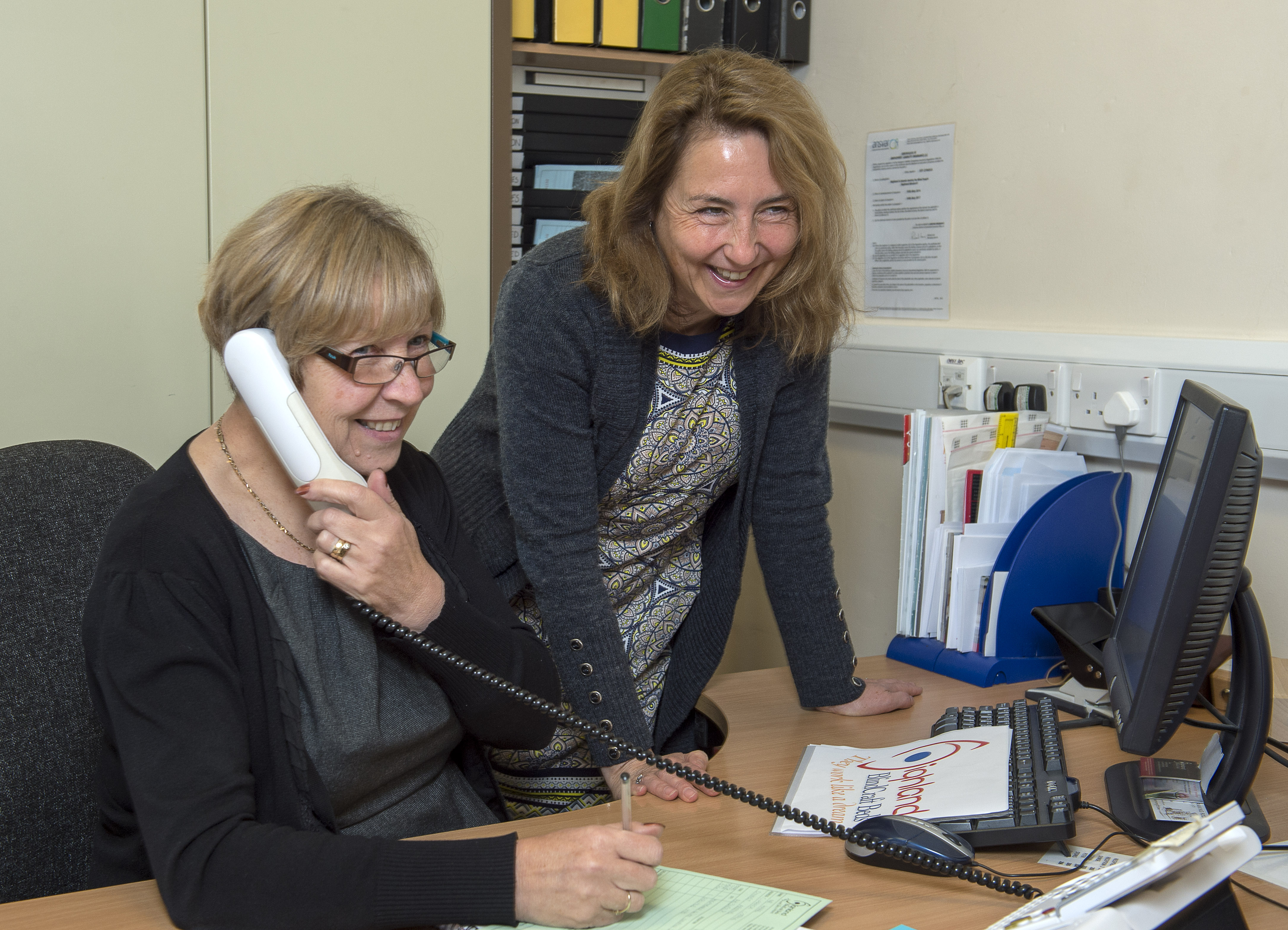 Two women at a desk - one standing, other on the phone