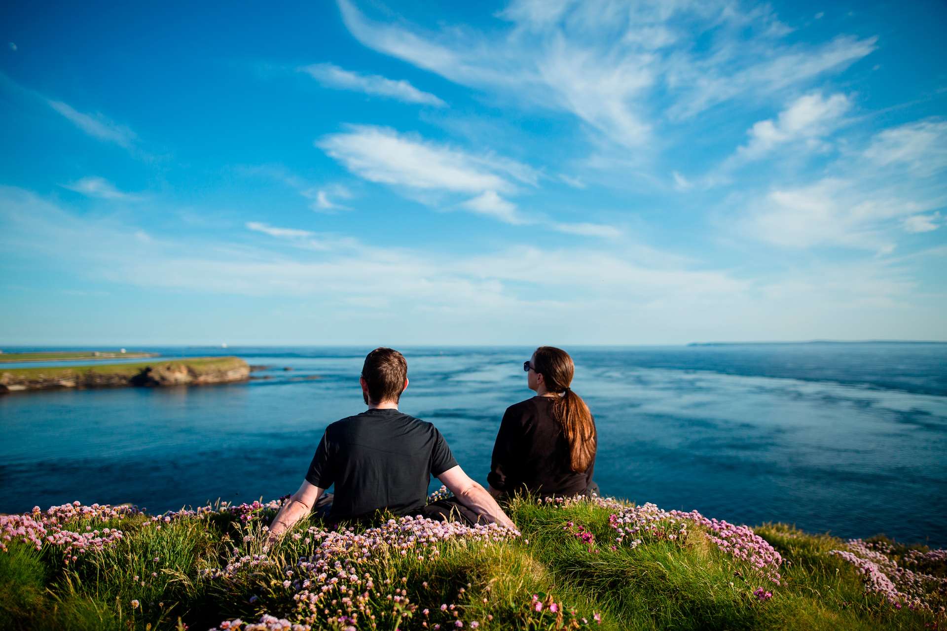 A couple sitting on top of flower covered hillside looking out to the bright blue ocean