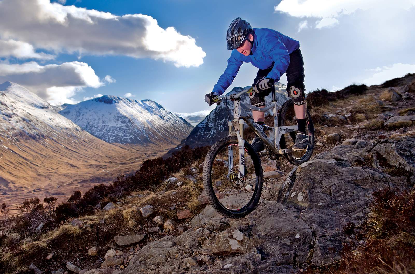 Person wearing blue jacket and black helmet on a bike perched downhill on black heather moorland